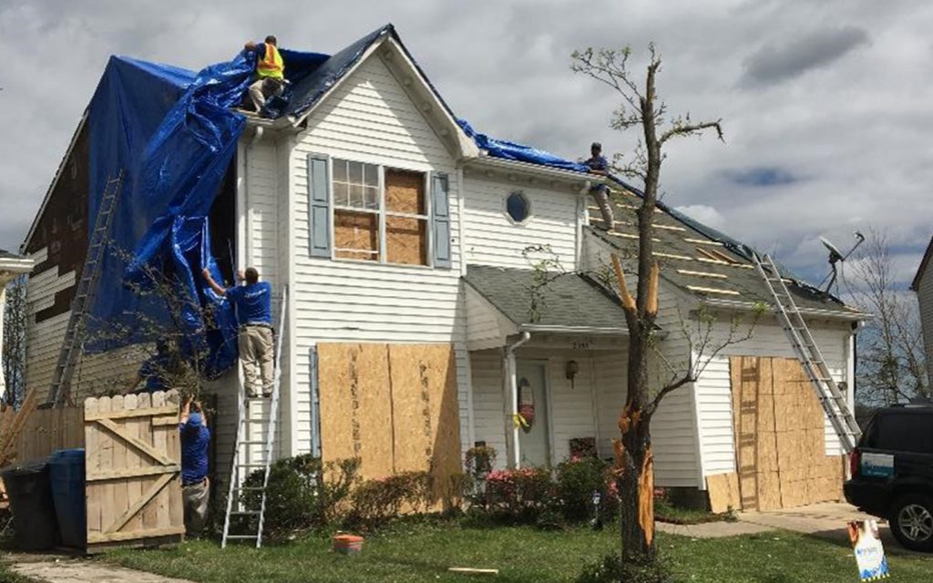 Emergency board-up crew securing a damaged house exterior with plywood and roof tarp